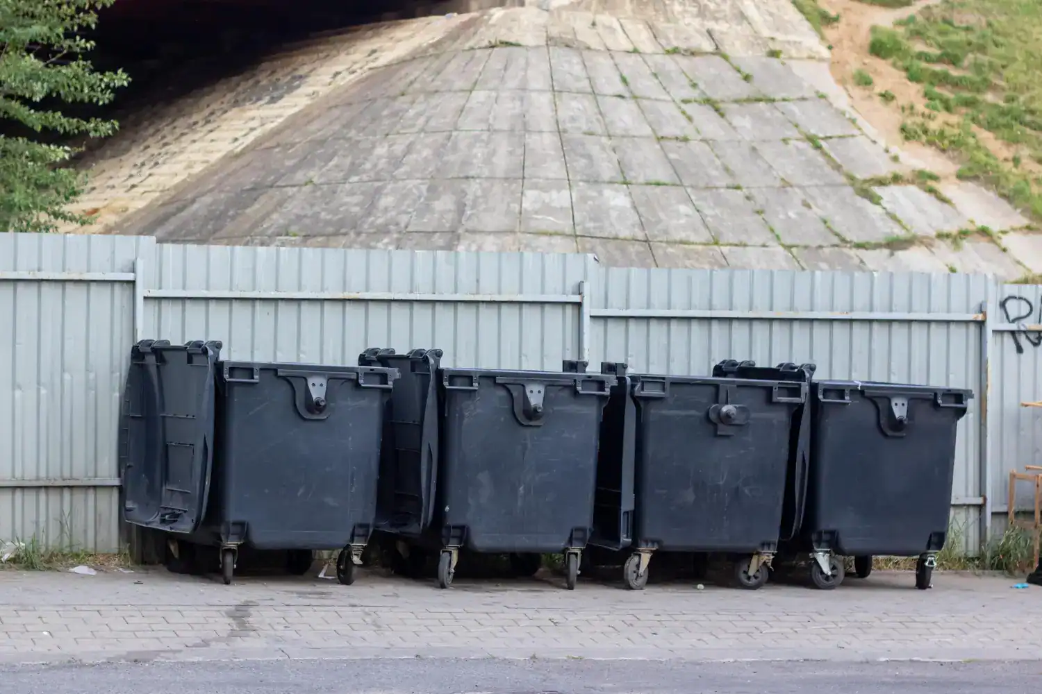 Multiple black commercial dumpsters lined up against a fence.