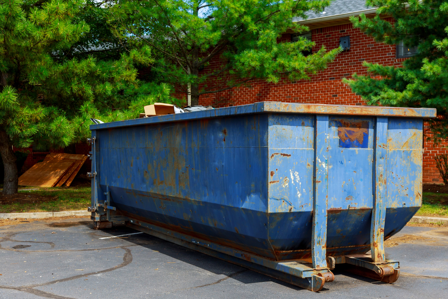 Rusty blue roll-off dumpster in a residential area, surrounded by trees and debris, illustrating effective waste management for home projects.
